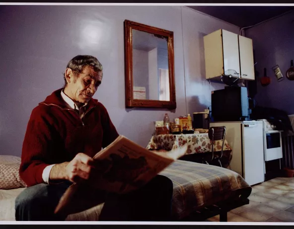 Homme âgé vu de face en biais, lisant son journal, miroir au mur. Guillaume Collanges, "Hôtels meublés - Si tu ne paies pas, tu t’en vas", entre 2000 et 2001, tirages gélatino-argentiques couleur, 41 cm x 62 cm, Collection du Musée national de l'histoire de l'immigration. 2007.59.4