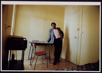 Homme avec moustache, se tenant debout et vu de face. Guillaume Collanges, "Hôtels meublés - Si tu ne paies pas, tu t’en vas", entre 2000 et 2001, tirages gélatino-argentiques couleur, 41 cm x 62 cm, Collection du Musée national de l'histoire de l'immigration. 2007.59.2