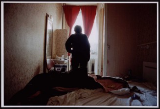 Silhouette de femme de dos derrière lit défait et devant fenêtre ornée de rideaux rouges. Guillaume Collanges, "Hôtels meublés - Si tu ne paies pas, tu t’en vas", entre 2000 et 2001, tirages gélatino-argentiques couleur, 41 cm x 62 cm, Collection du Musée national de l’histoire de l'immigration. 2007.59.6