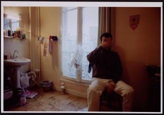 Homme vu de face se cachant le visage de sa main, devant coin salle de bains. Guillaume Collanges, "Hôtels meublés - Si tu ne paies pas, tu t’en vas", entre 2000 et 2001, tirages gélatino-argentiques couleur, 41 cm x 62 cm, Collection du Musée national de l’histoire de l'immigration. 2007.59.7 