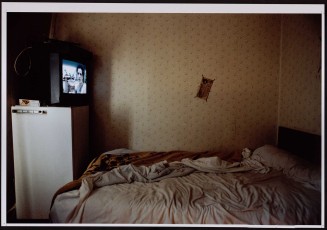 Vue sur un lit double défait devant poste de télé allumé. Guillaume Collanges, "Hôtels meublés - Si tu ne paies pas, tu t’en vas", entre 2000 et 2001, tirages gélatino-argentiques couleur, 41 cm x 62 cm, Collection du Musée national de l’histoire de l'immigration. 2007.59.8