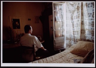 Homme vu de dos en biais devant rideau ensoleillé. Guillaume Collanges, "Hôtels meublés - Si tu ne paies pas, tu t’en vas", entre 2000 et 2001, tirages gélatino-argentiques couleur, 41 cm x 62 cm, Collection du Musée national de l’histoire de l'immigration. 2007.59.9