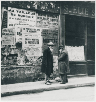 Deux hommes dans le Marais à Paris en avril 1933, devant une affiche pour un meeting de solidarité avec les juifs persécutés d'Allemagne. © Coll. Part./L. Karaïmsky, Cité nationale de l'histoire de l'immigration